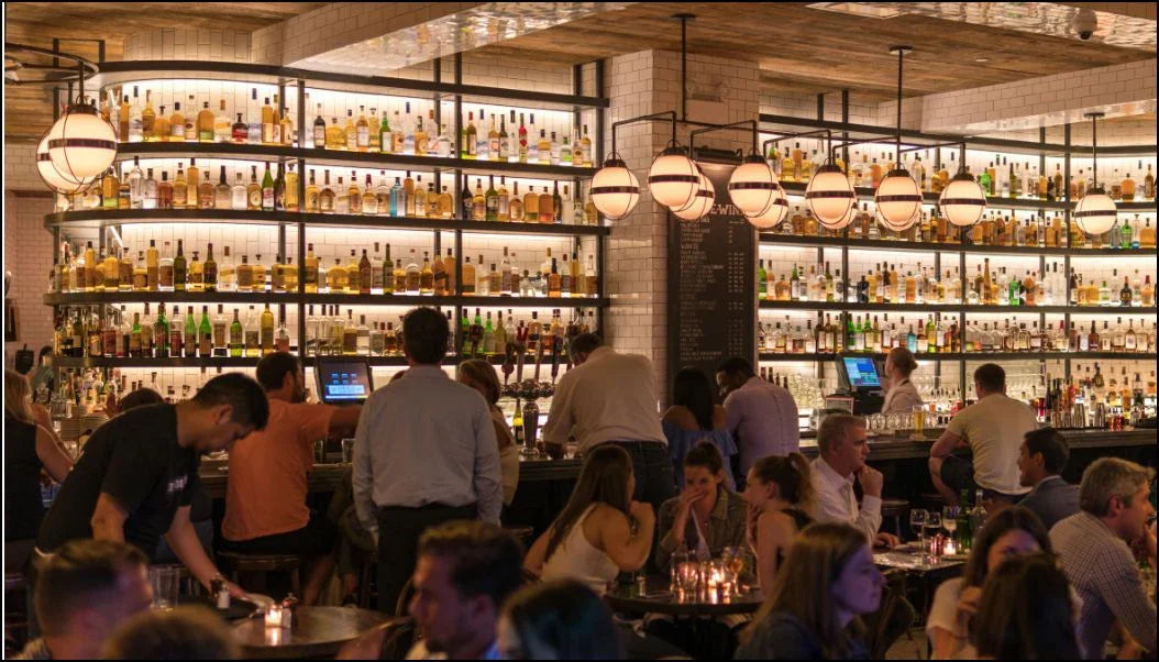Busy modern bar interior with patrons seated and standing, shelves filled with liquor bottles, and globe pendant lights
